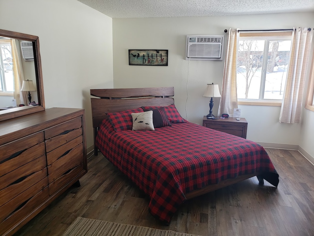A guest room features a spacious bed with a plaid red and black comforter. A wooden dresser and a mirror occupy one wall, while a window offers natural light and a view of the outdoors. An air conditioning unit is mounted above the bed.
