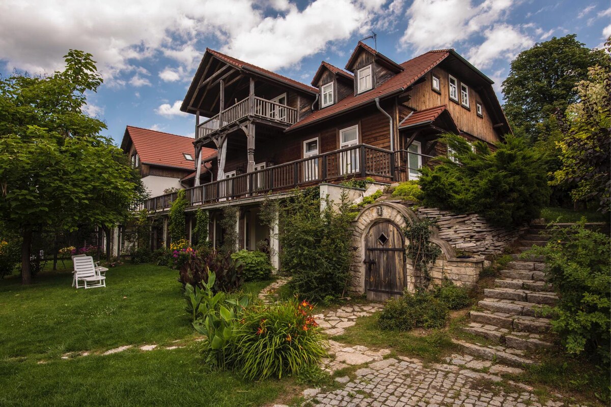 A charming multi-level building is set amidst lush greenery, featuring a welcoming porch with wooden railings. Pathways made of stone lead through the garden to an arched entrance, surrounded by colorful flower beds and well-maintained lawns. The sky displays an inviting blend of clouds.