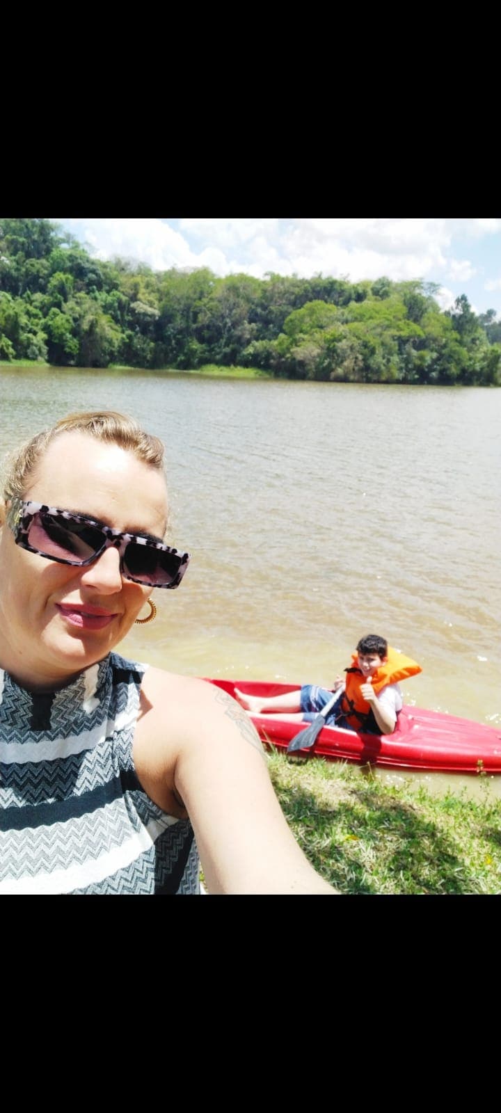 A calm lake is visible, bordered by lush green trees. A bright red kayak is positioned on the water, with a person seated inside wearing an orange life jacket. The shore is lined with grass, suggesting a peaceful outdoor setting.
