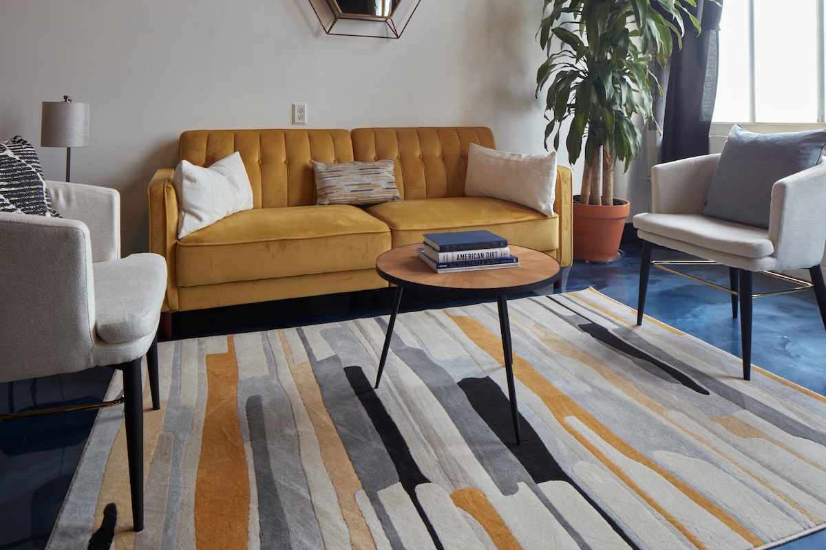 The living area features a yellow tufted sofa complemented by two light-colored armchairs. A round wooden coffee table sits on a striped area rug, adding texture and color to the space. A tall indoor plant and natural light from large windows enhance the room's ambiance.