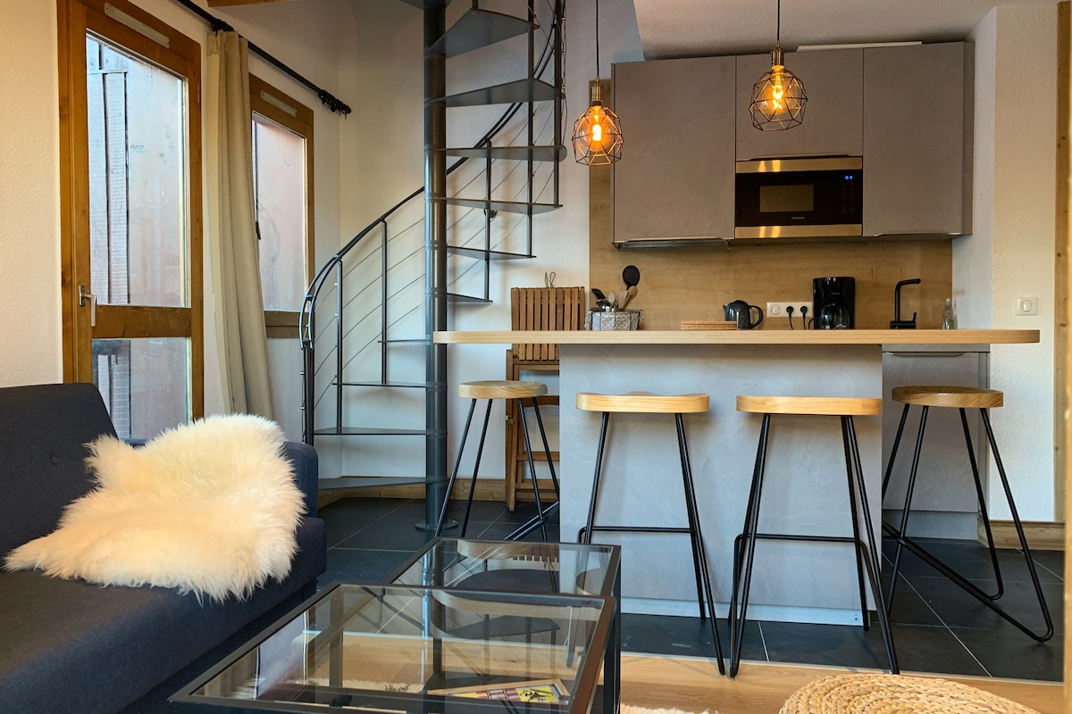 The open-plan living area features a modern kitchen with sleek cabinetry and a large breakfast bar. Two pendant lights hang above the bar stools, while a cozy seating arrangement includes a sofa with a soft throw. A glass coffee table complements the space.