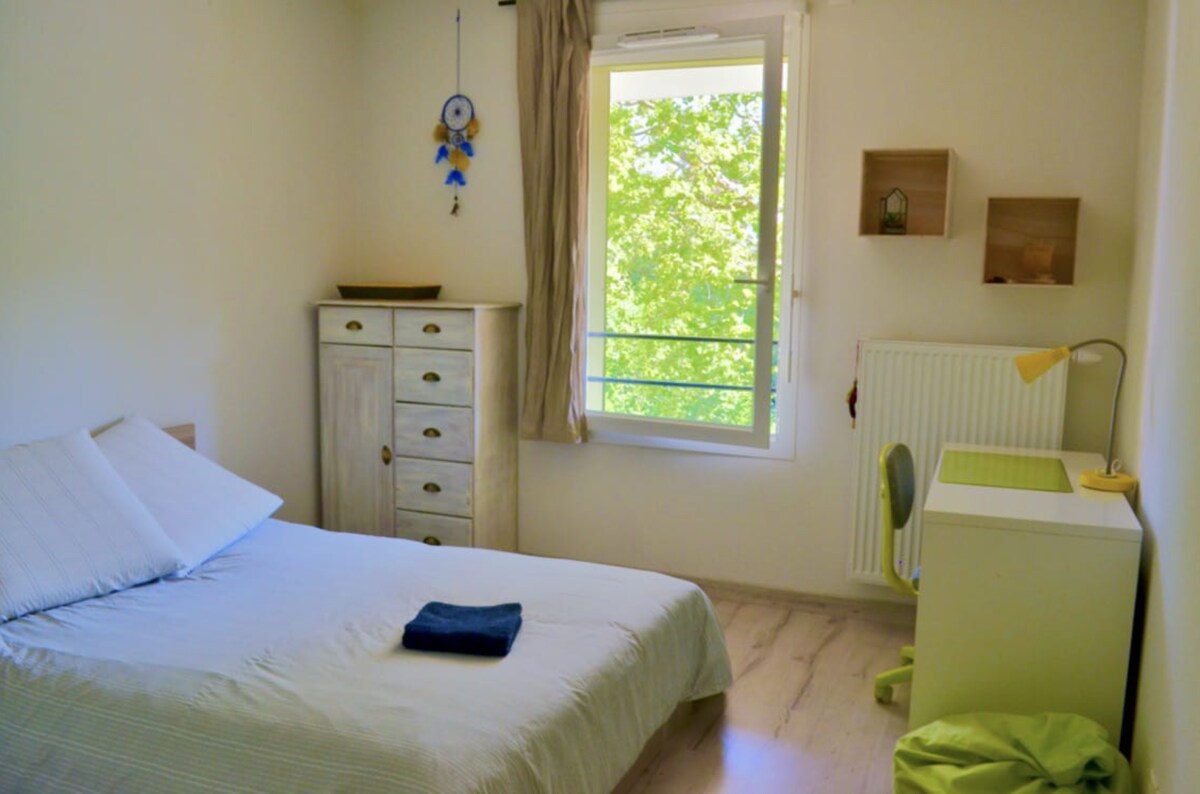 A bright bedroom features a spacious bed with white linens and a folded towel. A simple desk with a chair is positioned next to the window, which offers views of green trees outside. Wooden storage units add functionality to the serene space.