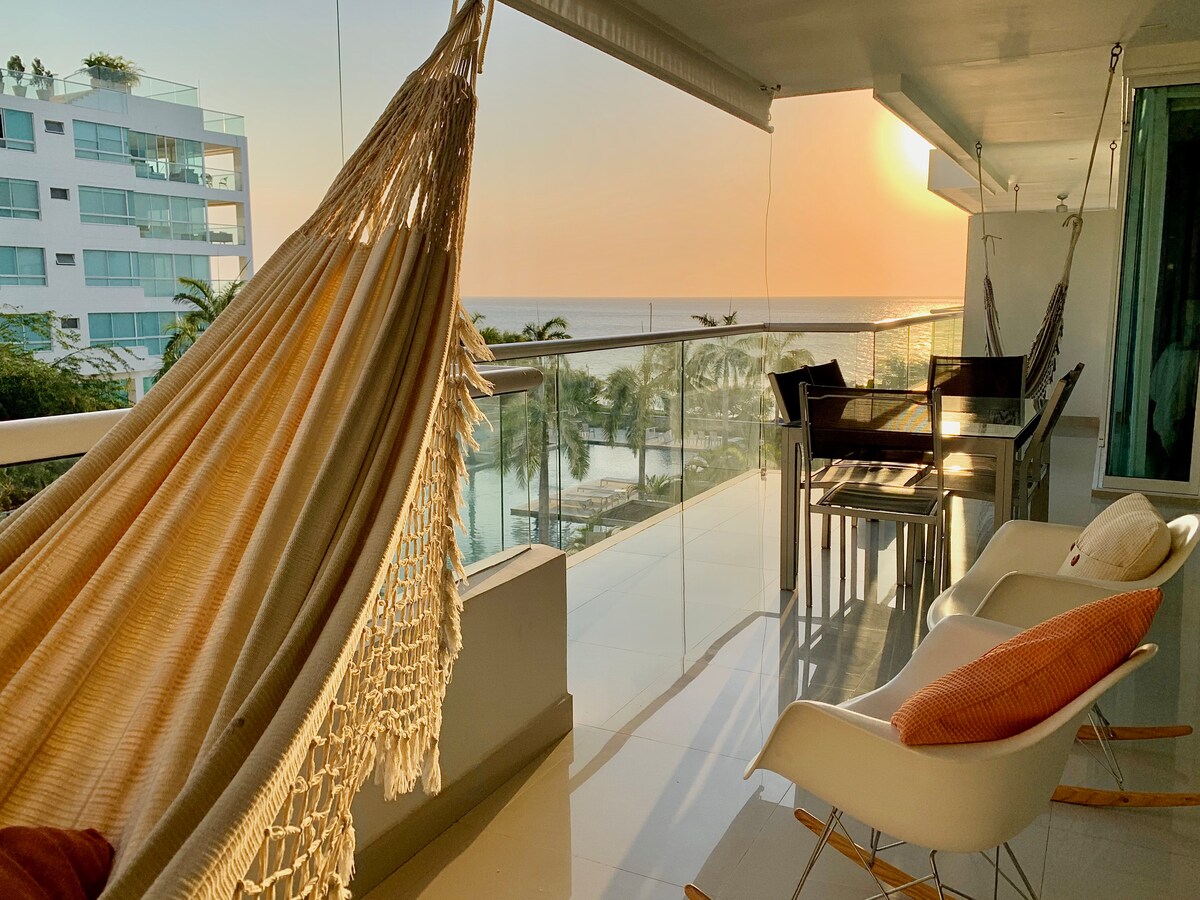 A balcony featuring a hammock and a small dining table, with chairs positioned for ocean views. The backdrop highlights a stunning sunset over the sea, while palm trees line the beach, creating a serene atmosphere.