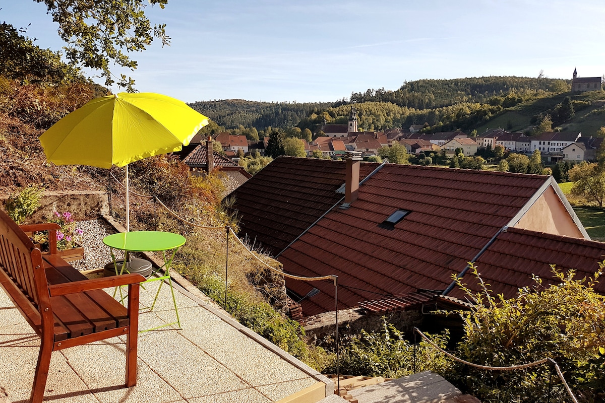 A small terrace is shown, featuring a wooden bench and a green table beneath a bright yellow umbrella. A picturesque view of rooftops and surrounding hills is visible in the background, creating a serene atmosphere.