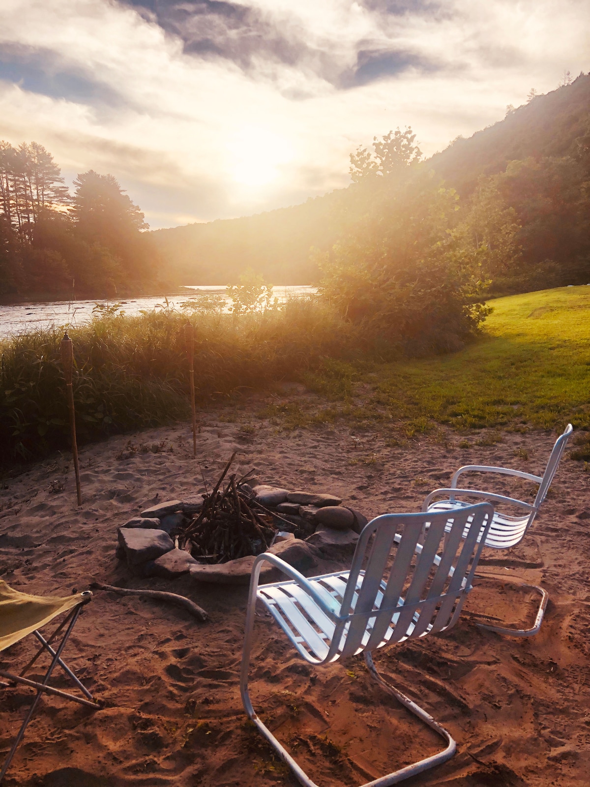 Seating is arranged around a stone fire pit on a sandy area, with two white chairs positioned toward the river. Sunlight filters through the trees as it sets behind distant mountains, illuminating the grassy landscape and creating a serene riverside atmosphere.