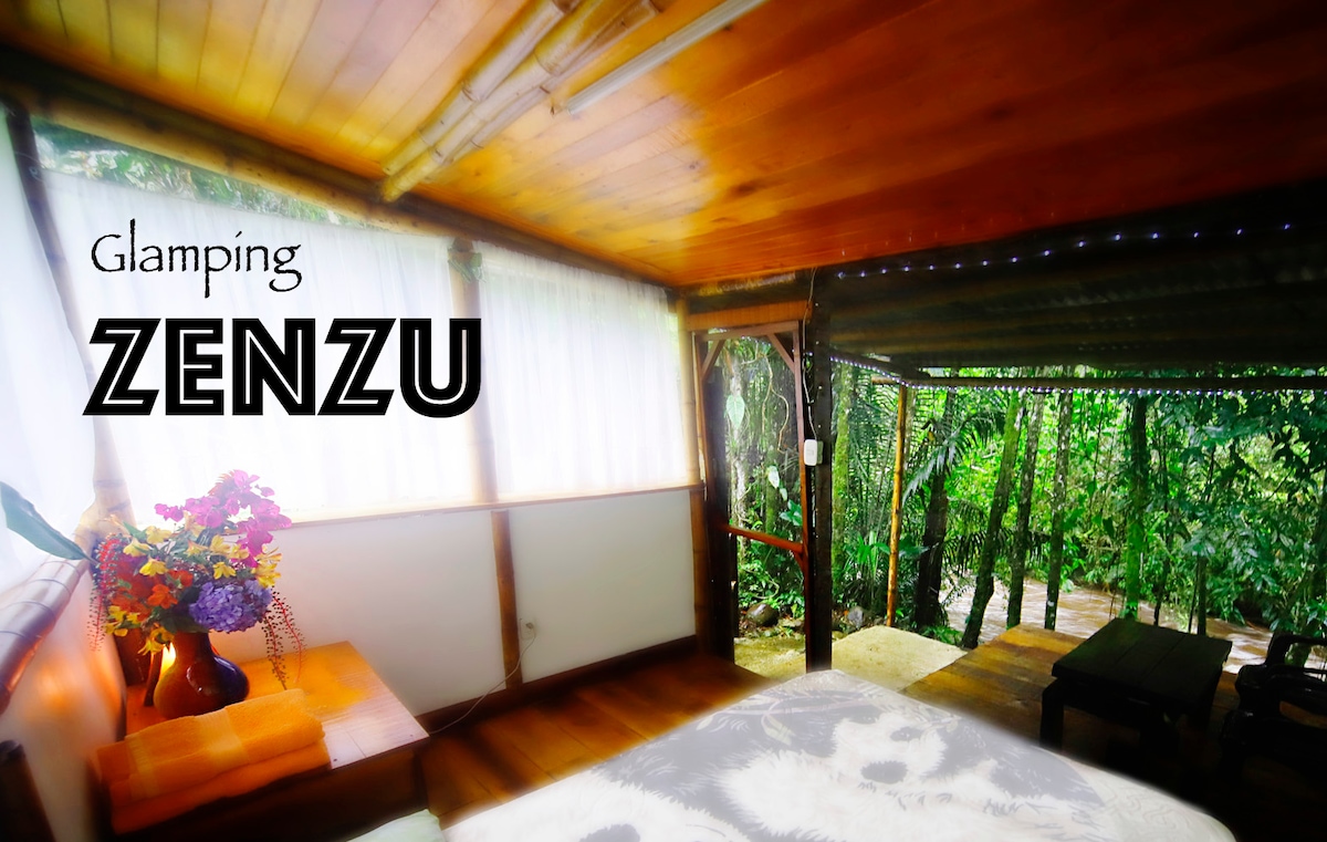 A glamping space named ZENZU features wooden walls and a sloped ceiling with warm lighting. Large windows provide views of the surrounding lush greenery. A bedside table supports a vase of colorful flowers, and a comfortable bed is positioned near the open space, inviting relaxation.