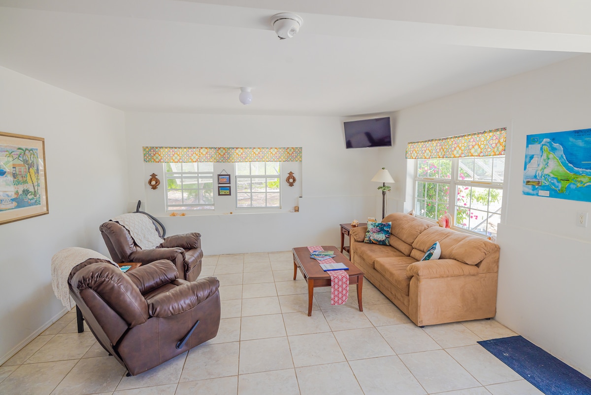 A spacious living area features a light-colored tile floor, complemented by large windows with colorful accents. Two comfortable recliner chairs are positioned alongside a beige sofa, with a wooden coffee table at the center. A flat-screen TV is mounted on the wall, providing entertainment options.
