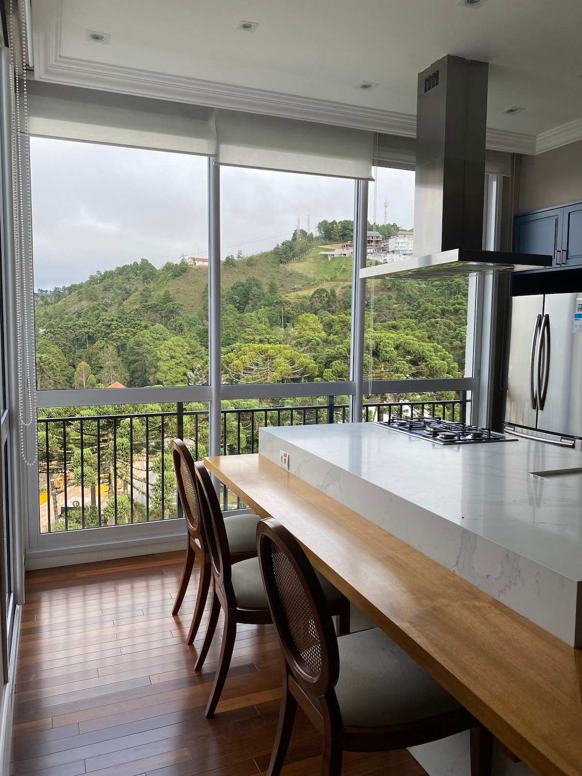 A modern kitchen showcases a spacious wooden dining table surrounded by four chairs. Large windows provide panoramic views of the lush mountains, allowing natural light to fill the room. A sleek stainless steel range hood is mounted above the cooktop, combining functionality with design.