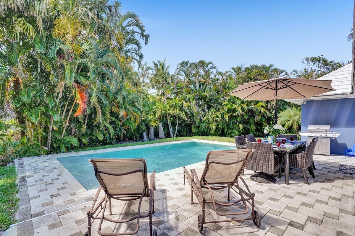 An expansive outdoor area features a private heated pool surrounded by lush tropical foliage. Two lounge chairs are positioned nearby on a patterned stone patio, alongside a dining table with an umbrella. A grill is visible in the background, enhancing the outdoor space.