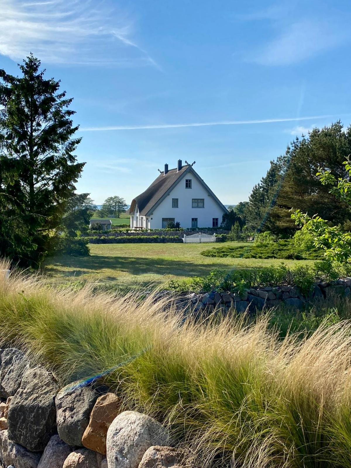 A charming double house is set against a clear blue sky, featuring a thatched roof and landscaped grounds. Lush greenery surrounds the property, with a stone wall in the foreground. The spacious yard includes shrubs and trees, providing a serene environment.