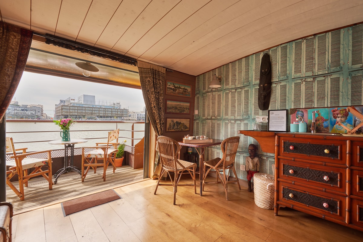 An inviting living space features a wooden floor and a cozy dining area with a round table surrounded by four chairs. Large glass doors open to a balcony overlooking the water, allowing natural light to fill the room. Vintage decor adds character to the space.