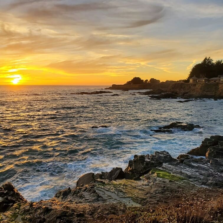 A coastal scene at sunset showcases vibrant orange and yellow hues against the horizon. Waves gently crash against rocky outcrops, while silhouettes of nearby cliffs and homes provide a natural framing. The sky features soft clouds, reflecting the day's last light.
