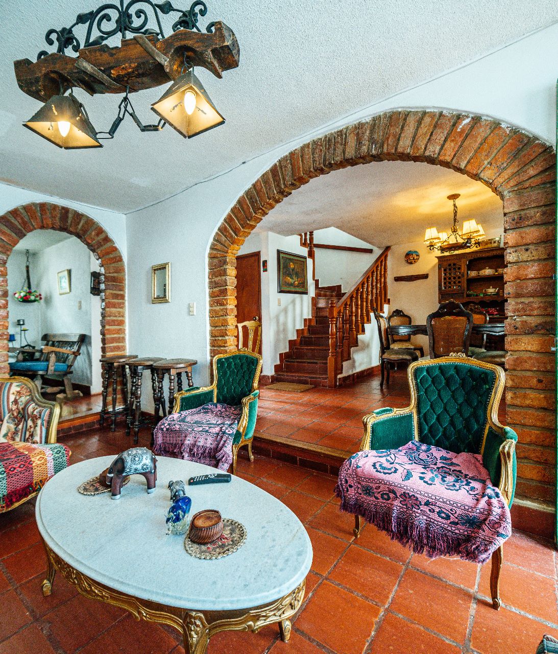 A welcoming living area is highlighted by architectural arches and terracotta tiled flooring. Two green upholstered chairs with intricate patterns flank a marble coffee table. A wooden staircase leads to the upper floor, while various decorative elements add character to the space.