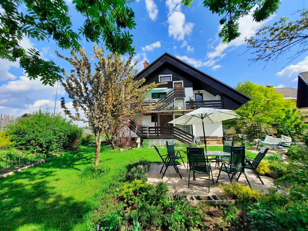 The exterior of the villa is surrounded by lush greenery and features a spacious garden. A patio area with multiple lounge chairs and an umbrella invites relaxation. The large balcony is visible on the second floor, complemented by a backdrop of vibrant blue skies and scattered clouds.