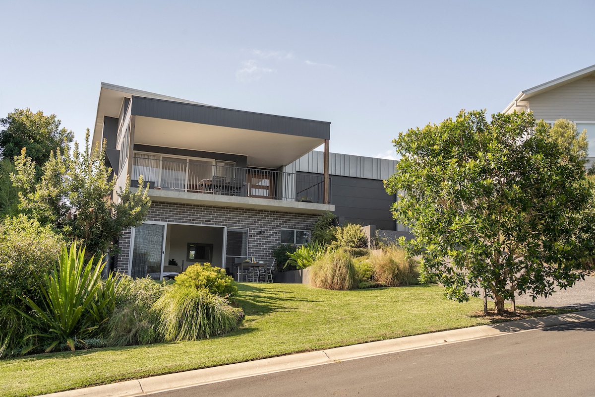 The modern home is situated on a gently sloping lawn, featuring lush greenery and landscaped gardens. A spacious upper balcony is visible, framed by large windows on the lower level, creating a welcoming connection with the outdoors.