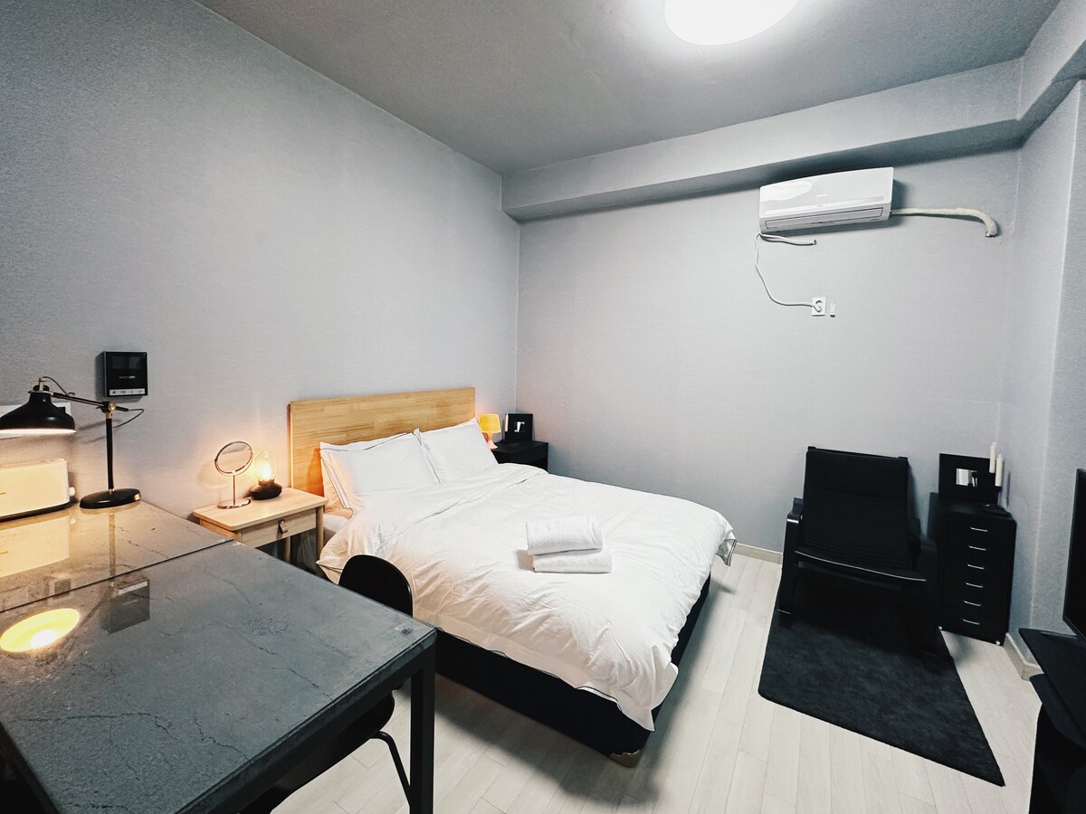 A spacious studio is shown with a queen size bed featuring crisp bedding. A sleek desk is visible, alongside a comfortable chair. The walls are painted in a soft grey, and a minimalist black accent rug complements the modern decor. An air conditioning unit is mounted on the wall.