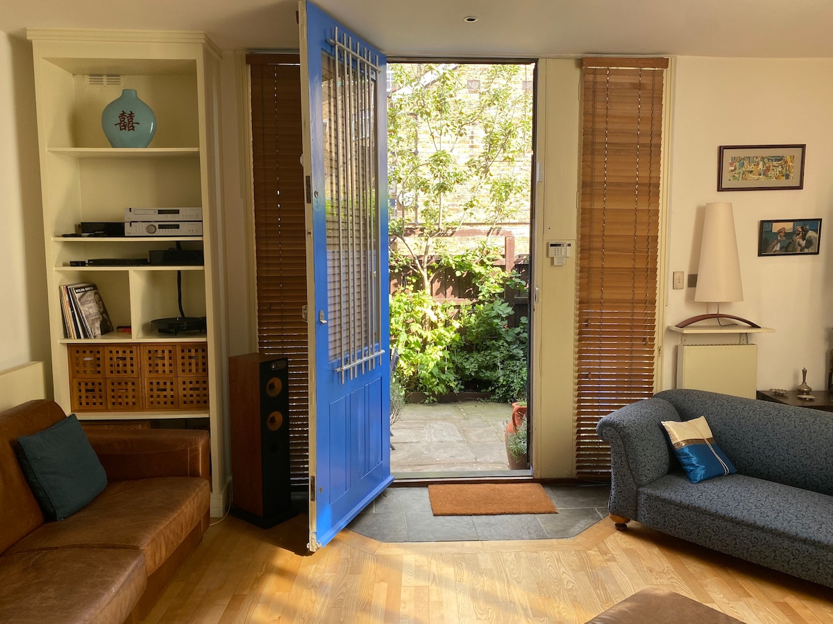 A bright living area is showcased, with a blue front door open to reveal a small garden. Wood flooring extends throughout, complemented by a light-colored sofa and a brown seating area. Natural light filters in through the wooden blinds, enhancing the inviting atmosphere.