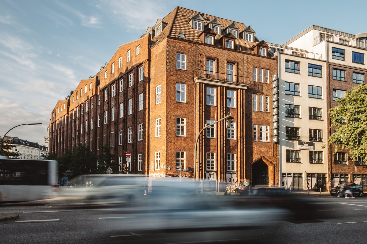A brick building with a distinctive architectural style stands prominently against a clear sky. Large windows reflect the surroundings, while a street in front shows blurred movement of passing vehicles, conveying a lively urban atmosphere.