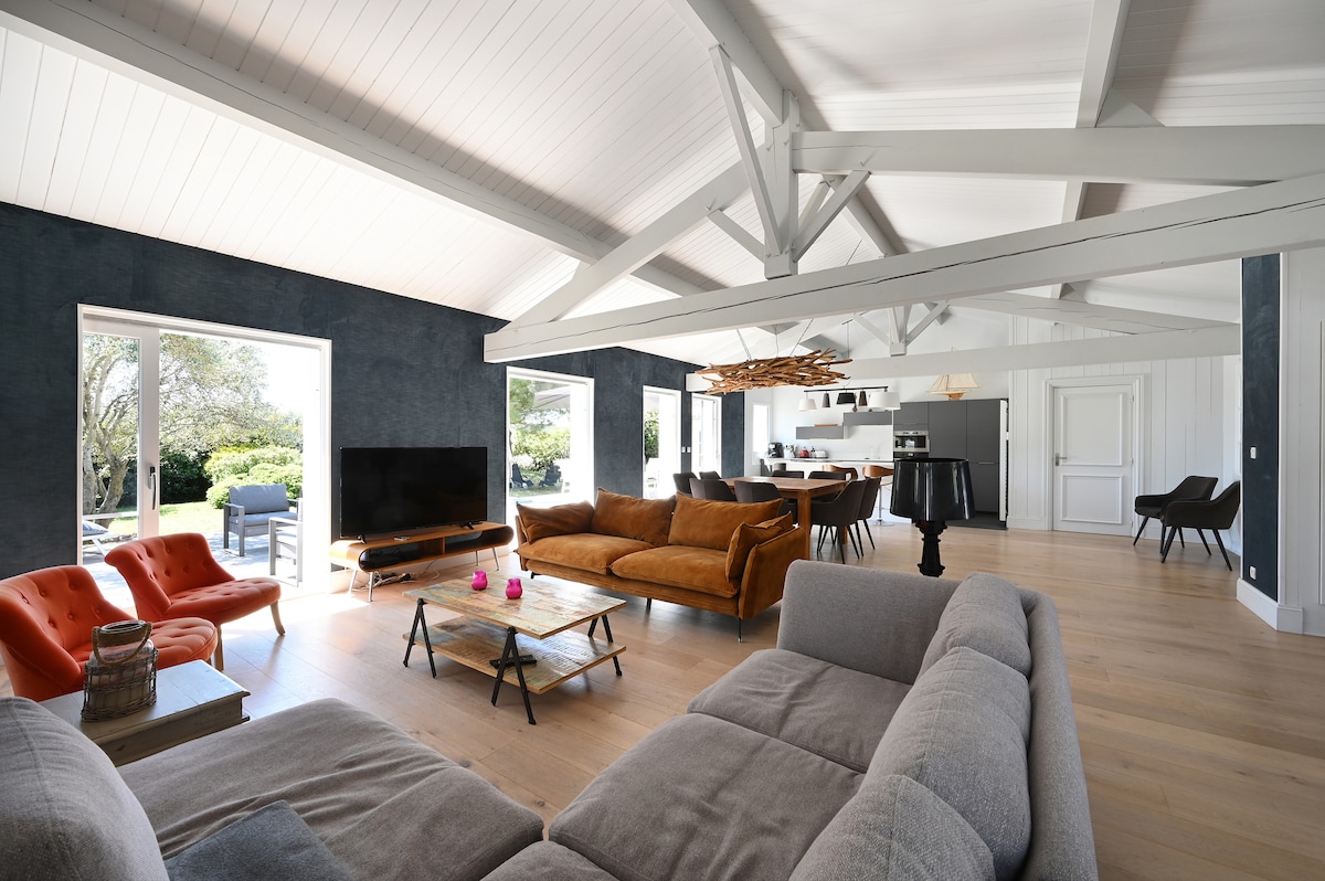 An open living space features large windows that invite natural light and provide views of the garden. Comfortable seating options include a spacious grey sofa and vibrant orange chairs. A wooden coffee table sits centrally in the room, complementing the modern decor.