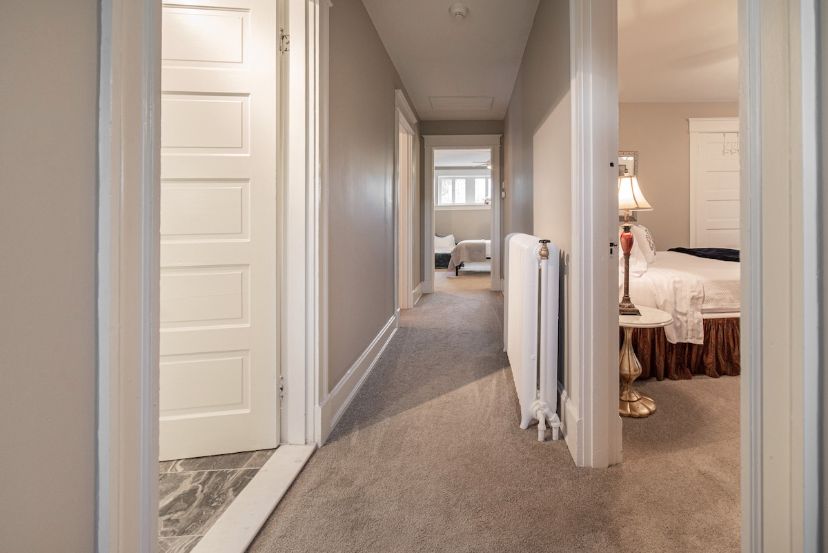 A well-lit hallway is seen, featuring plush carpeting and neutral-toned walls. Several doors lead to different rooms, with a glimpse of a bedroom at the end of the corridor. Natural light enters from the windows, creating a welcoming ambiance.