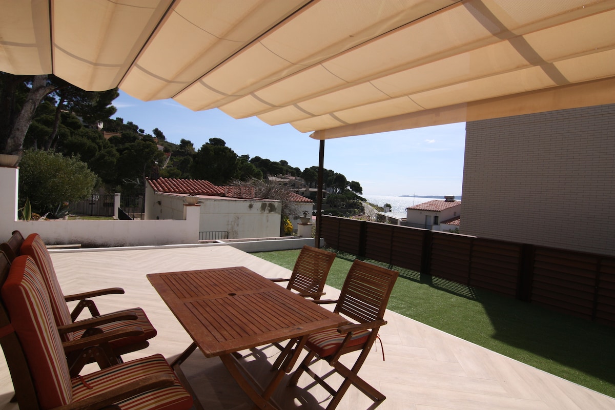 A spacious private patio is shown with a wooden table surrounded by slatted chairs. A retractable awning provides shade, while the distant view of the sea and tree-lined landscape is visible beyond the patio's edge. The lawn area is neatly maintained.