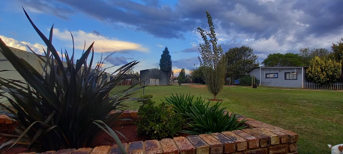 A spacious garden area features well-maintained green grass and a variety of plants. In the background, simple buildings are visible, surrounded by trees and a clear sky. The landscape creates a tranquil atmosphere, ideal for relaxation.