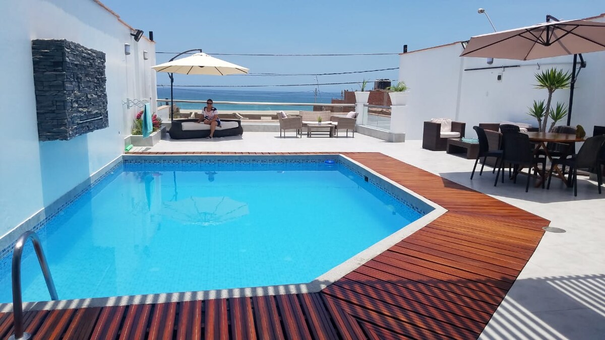 A sparkling blue pool is framed by a wood deck and surrounded by shaded lounge areas. Umbrellas provide shade for seating arrangements, while ocean views are visible in the background, enhancing the outdoor space's relaxation potential.