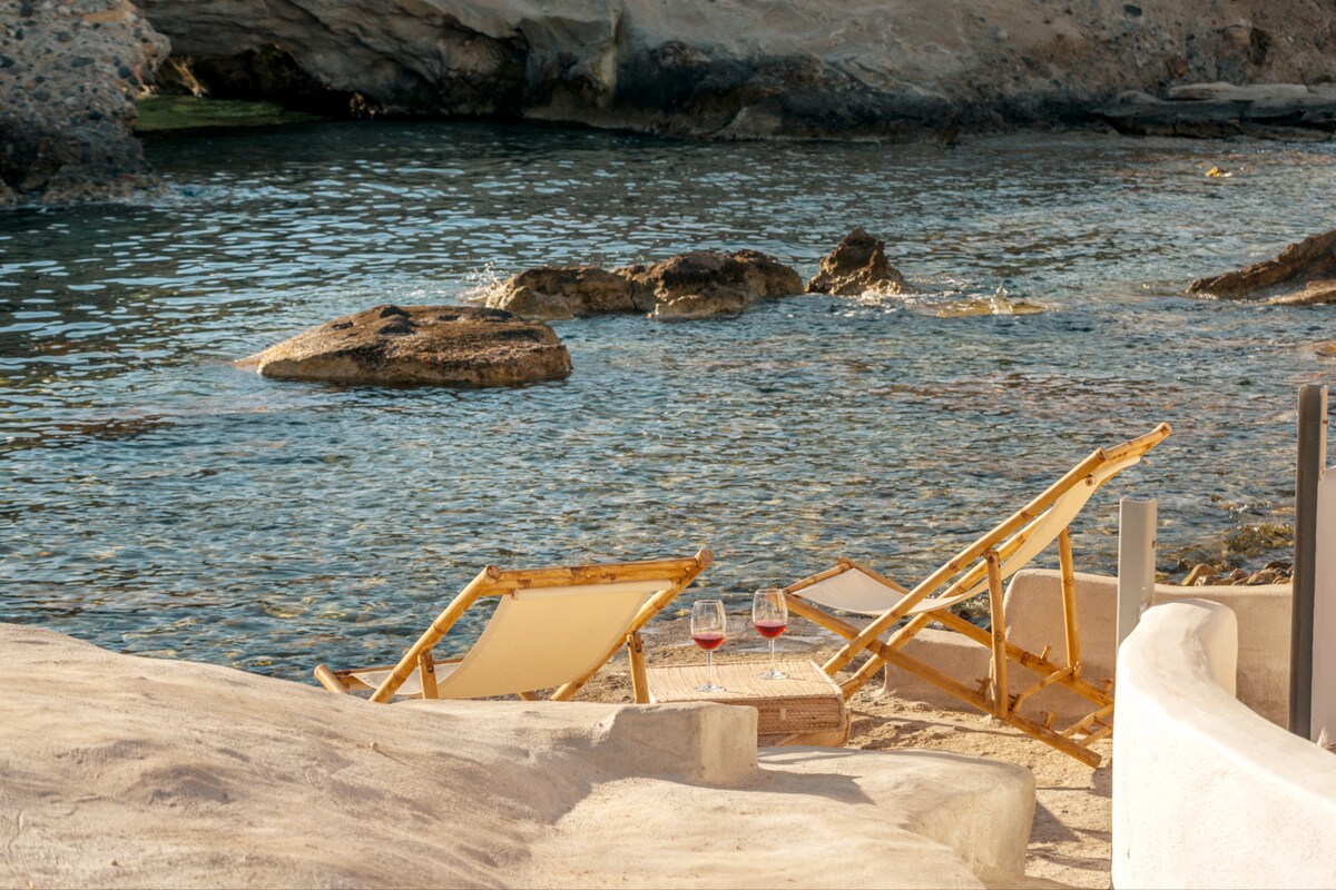 Two wooden lounge chairs are positioned on a sandy beach, facing calm waters with a rocky shoreline. A small table holds two glasses of red wine, reflecting soft sunlight. The scene is peaceful, with gentle waves lapping at the shore.