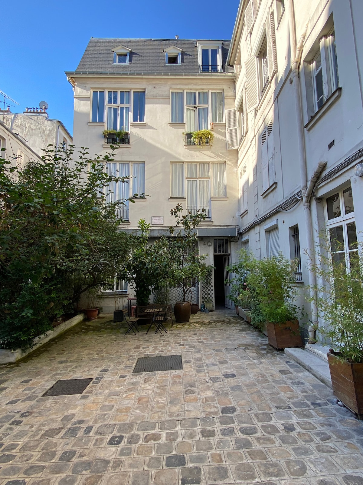 A tranquil courtyard is surrounded by buildings, showcasing a cobblestone pathway lined with greenery. Tall windows with shutters add charm to the facade, while potted plants and a small seating area invite relaxation in the serene outdoor space.