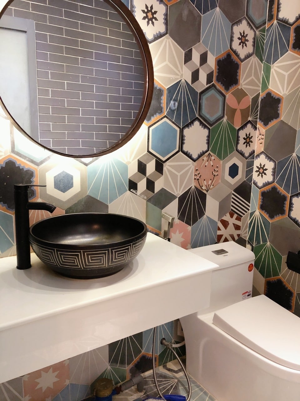 A modern bathroom features a striking hexagonal-patterned wall. A sleek, dark bowl sink is set on a minimalist white countertop, complemented by a round mirror above. A compact toilet sits adjacent, completing the contemporary design.