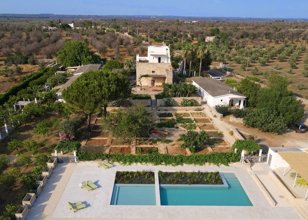 An aerial view displays the expansive grounds of the Masseria Gianferrante, featuring a serene swimming pool bordered by lush greenery. The surrounding gardens include pathways and various plantings, while the rustic stone structure of the Masseria is visible in the background.