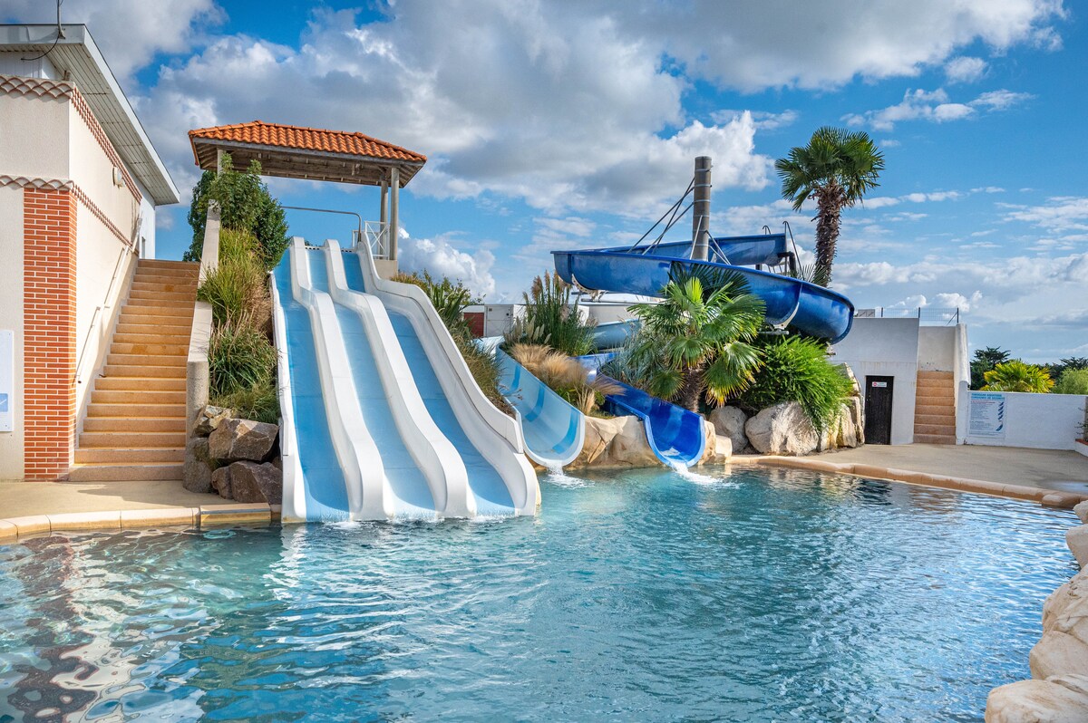 A water park area features two white and blue water slides leading into a pool surrounded by palm trees and vibrant foliage. The scene is bright, with fluffy clouds above and a patio area visible in the background, providing a welcoming environment for relaxation and play.