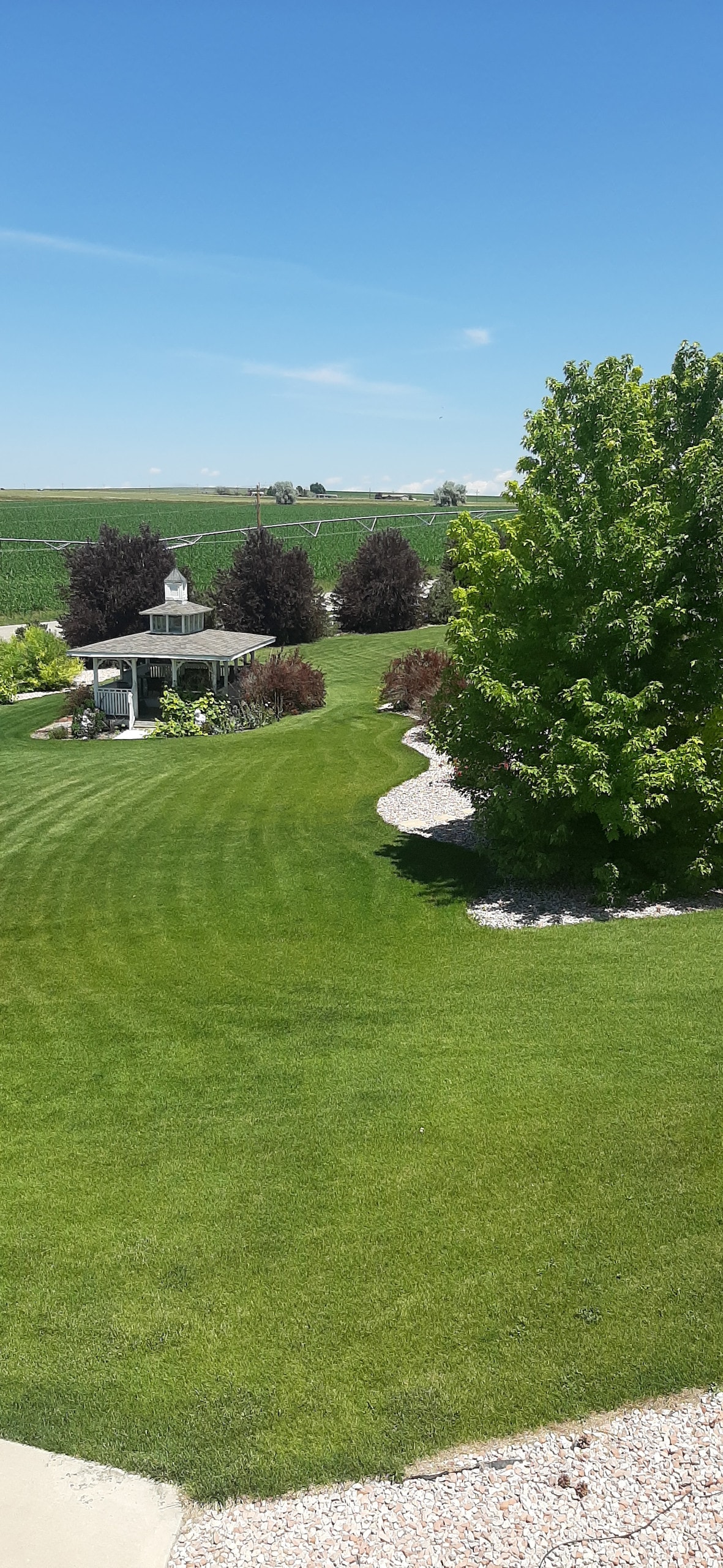 A well-maintained yard with lush green grass and landscaped shrubs is depicted. A gazebo is visible in the distance, surrounded by flowering plants. The scene is complemented by a winding gravel path that guides the eye through the open space under a clear blue sky.