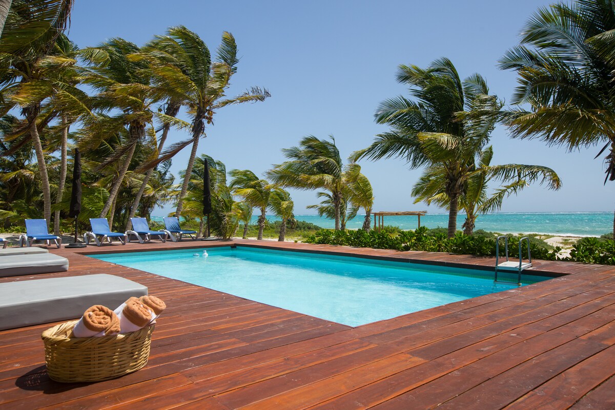 A serene pool area features a refreshing swimming pool surrounded by wooden decking. Sun loungers are arranged nearby, with palm trees swaying in the breeze. The turquoise waters of the Caribbean Sea and sandy beach are visible in the background.