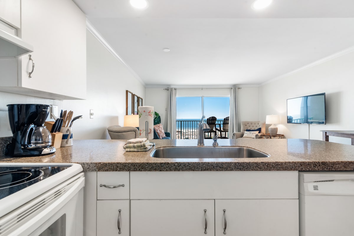 A view of the kitchen area features white cabinetry and a granite countertop. An electric stove, sink, and various kitchen utensils are visible. The open layout connects to a cozy living space with soft seating, illuminated by natural light from a large sliding glass door leading to the ocean view.