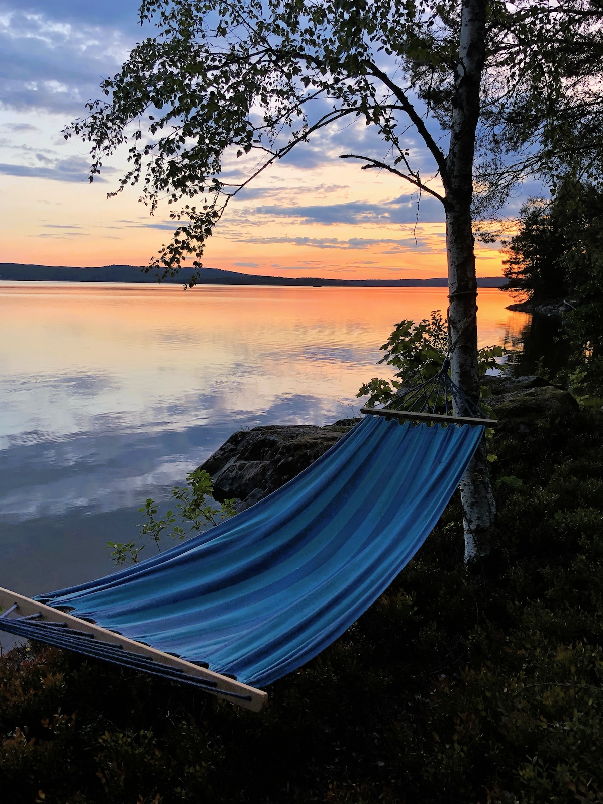 A blue-striped hammock is suspended between two trees, overlooking a calm lake at sunset. The tranquil water reflects the soft hues of orange and purple from the sky. Lush greenery frames the scene, providing a serene outdoor setting.