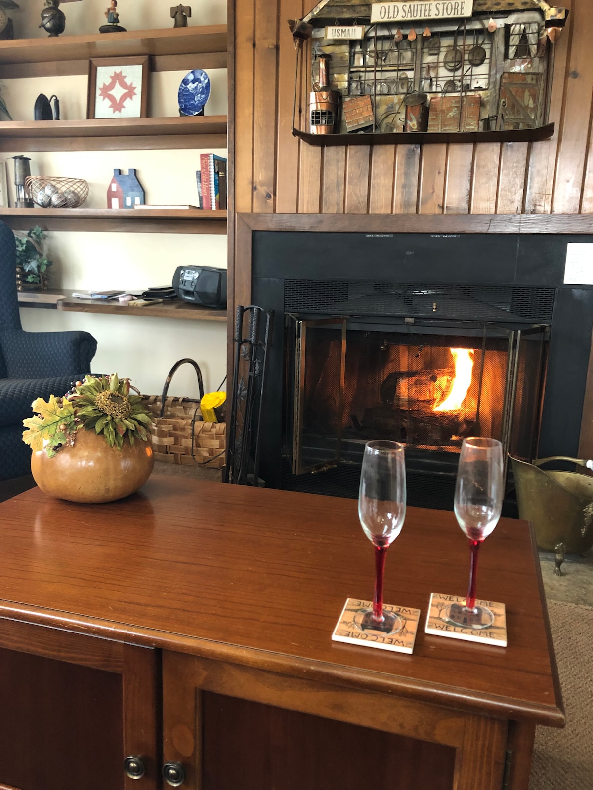 A cozy living area features a wooden coffee table topped with two champagne flutes and coasters. A warm fire flickers in the fireplace, surrounded by wood paneling and decorative shelving with various items. A comfortable armchair is visible in the background.