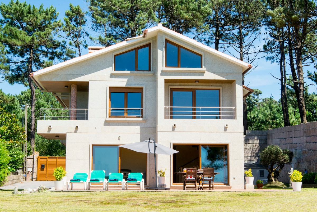 The villa's exterior is showcased with large windows that offer ample natural light. A patio area features sun loungers and a dining set under a parasol. Surrounding tall trees contribute to a serene setting, creating a space for relaxation.