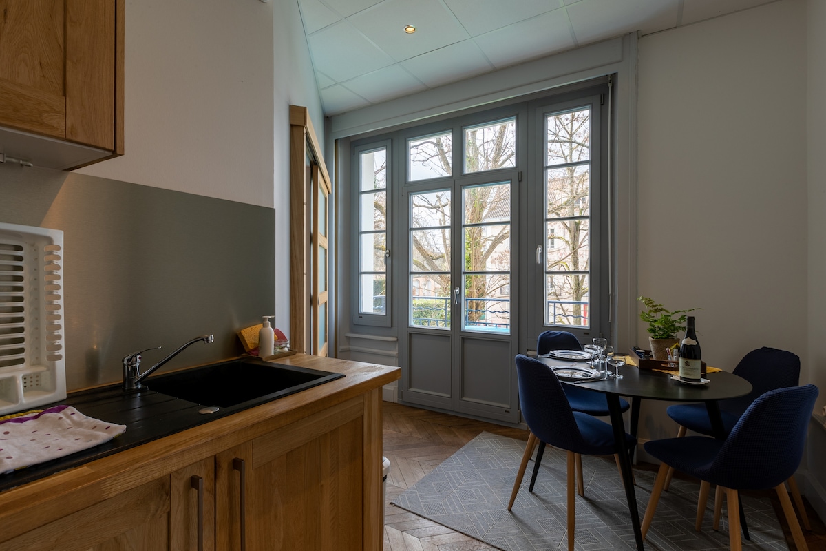 A dining area is observed with a round table and four blue upholstered chairs. Natural light filters through large windows that provide views of trees outside. The kitchen features wooden cabinetry and a black sink, complemented by a light-toned area rug on the floor.