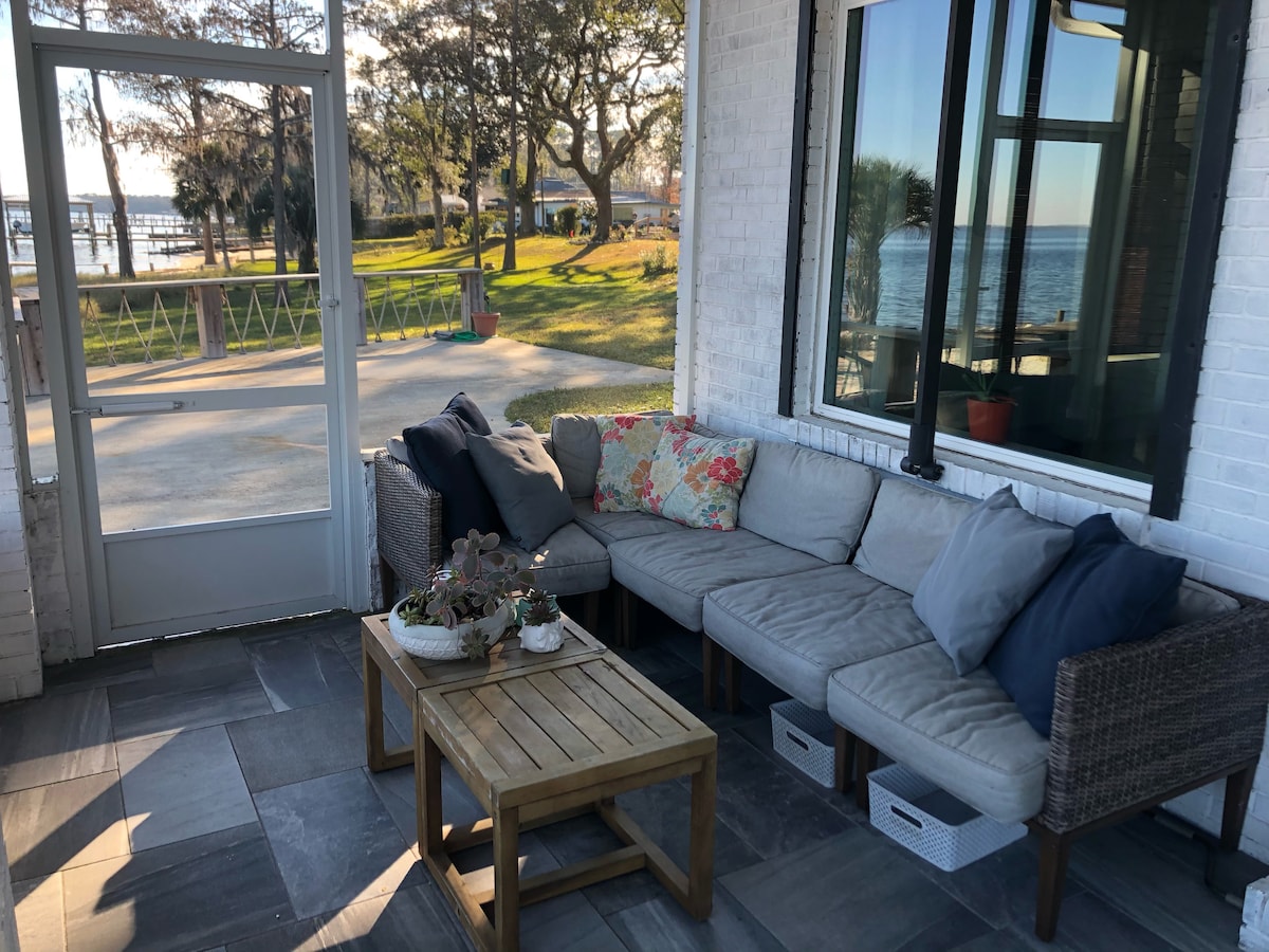 A screened-in porch features a comfortable seating arrangement with plush cushions in neutral tones. A wooden coffee table sits in the center, complemented by a potted plant. The backdrop reveals views of the waterfront and outdoor greenery, providing a peaceful setting.