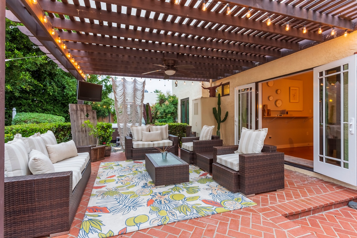 An outdoor living space is showcased under a wooden pergola with market lights. Comfortable seating arrangements with soft cushions surround a glass-top coffee table. A vibrant area rug adds color, and the background features lush greenery and an outdoor television.