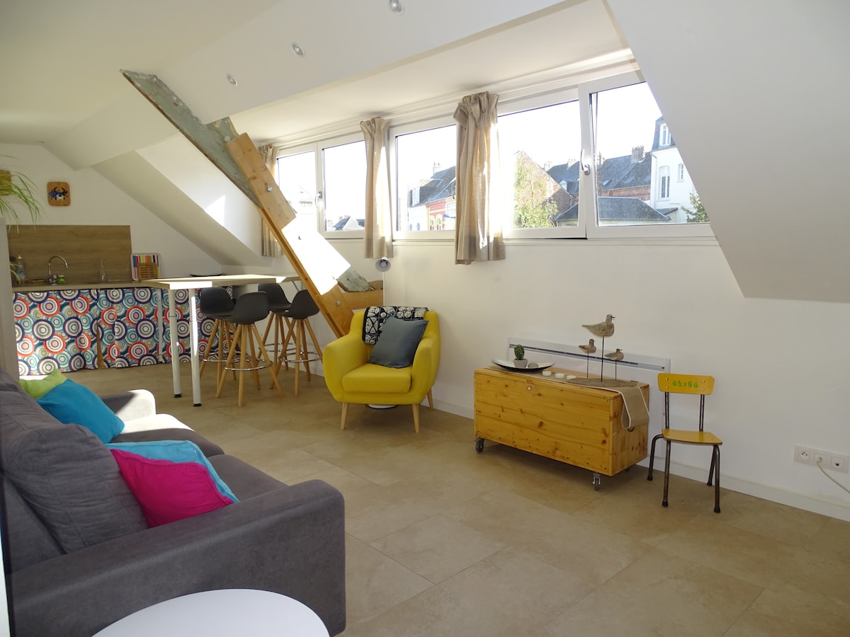 A bright living space is presented, featuring a comfortable sofa adorned with colorful cushions. A small wooden table and chairs are arranged beside a kitchenette, which is decorated with a patterned backsplash. Large windows allow natural light to enter, creating an airy atmosphere.