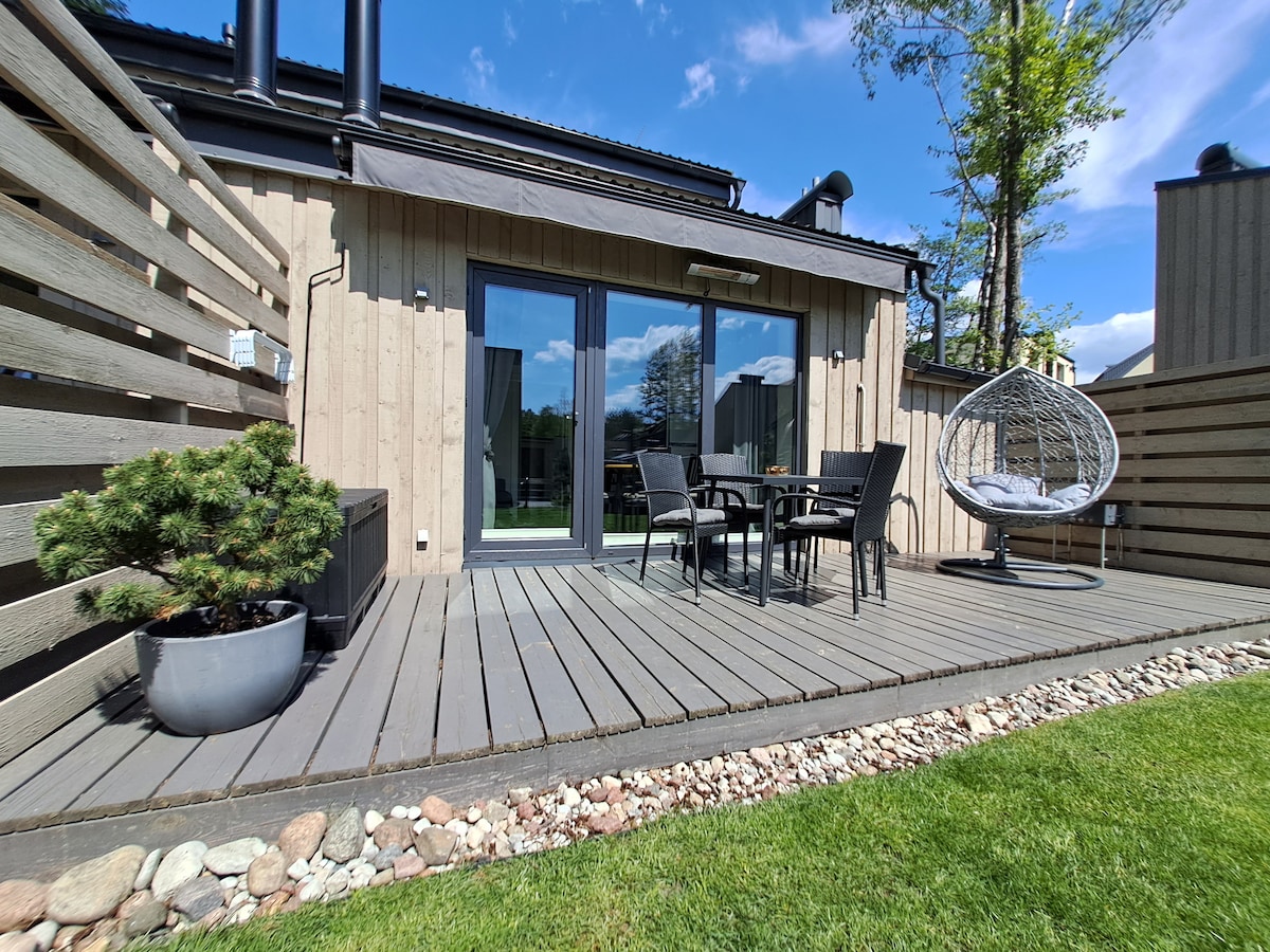 A private terrace features wooden decking surrounded by a verdant lawn. Black outdoor furniture, including a table and four chairs, is set for relaxation. A stylish hanging chair provides additional seating, with a small potted tree enhancing the space.