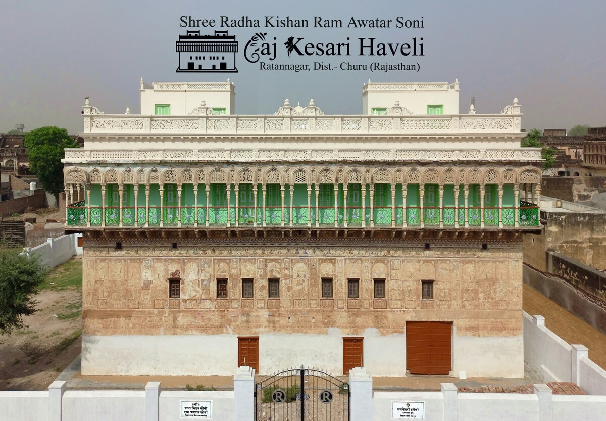 The exterior of Gaj Kesari Haveli is displayed, featuring intricate wall frescoes and a symmetrical façade. Ornate balconies with green shutters line the upper level, and smooth white walls create a striking contrast against the light brown of the frescoes. The surrounding area is enclosed by a low wall.