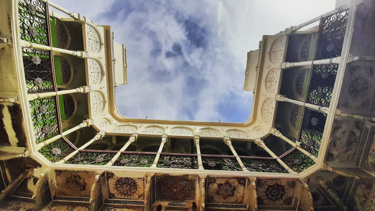 A view from below showcases an intricately designed courtyard surrounded by ornate balconies. The architecture features elaborate frescoes and decorative lattice work, contrasting against the cloudy blue sky. The structural symmetry and craftsmanship of the haveli are highlighted from this unique perspective.