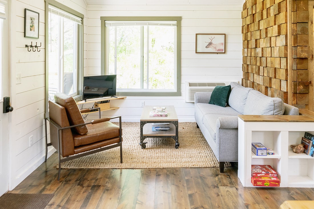 A cozy living area is highlighted by natural light from large windows. A gray sofa and a brown leather armchair are positioned around a central coffee table. A wood accent wall adds texture, while a TV is mounted on the opposite wall. Board games are visible on a shelf.