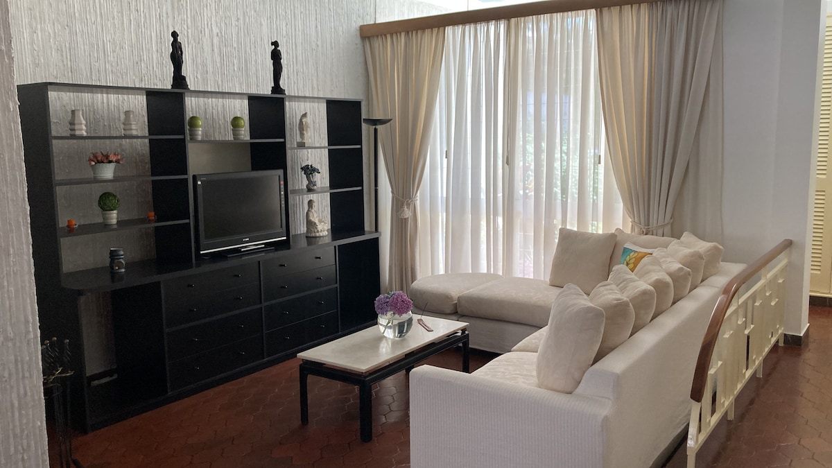 The living area displays a comfortable sectional sofa and a chic coffee table, complemented by a dark media console featuring decorative accents. Sheer curtains allow natural light to flow in, enhancing the openness of the space.
