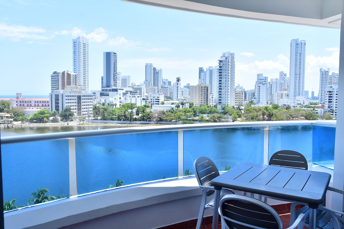 A balcony offers a panoramic view of the city skyline, featuring tall buildings beside a tranquil body of water. A small table and chairs are positioned for relaxation, creating a peaceful outdoor space surrounded by the vibrant landscape.