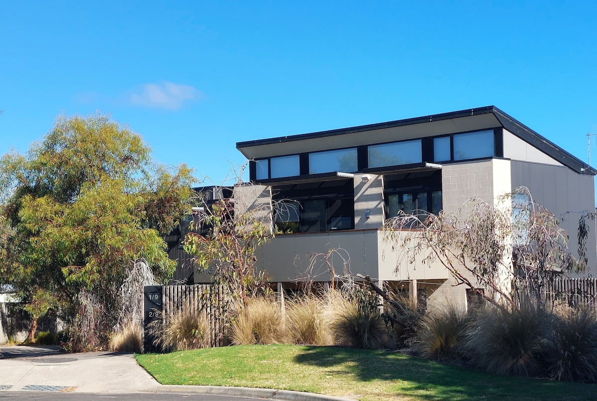 The contemporary townhouse is depicted surrounded by natural landscaping, featuring a mix of trees and shrubs. The structure has large windows and a modern architectural design, complemented by a clear blue sky overhead.
