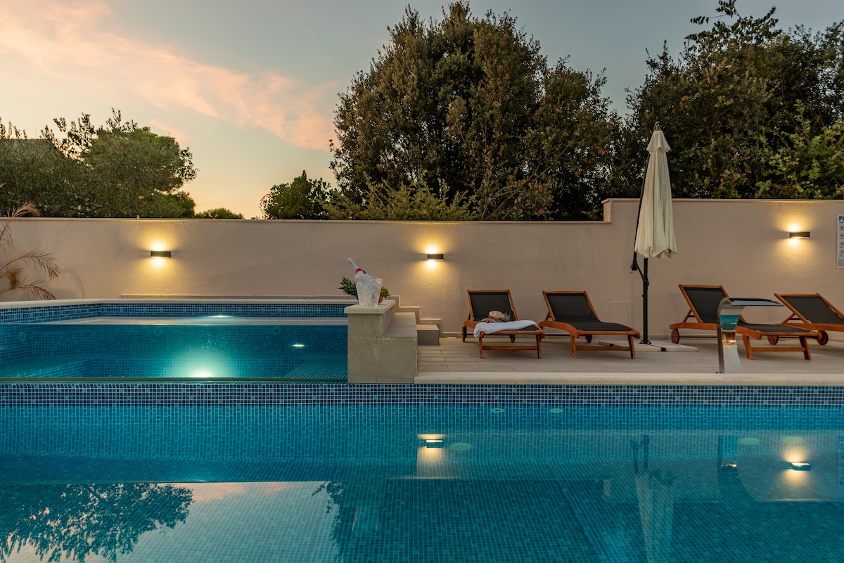 A tranquil outdoor area features a heated pool with a shimmering blue surface. Sun loungers are arranged beside the pool, bordered by greenery. Soft lighting from wall sconces provides illumination as dusk approaches, enhancing the serene setting.